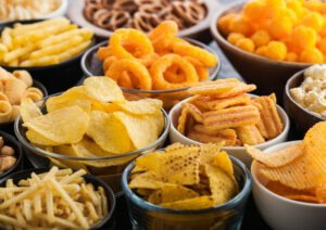 All classic potato snacks with peanuts, popcorn and onion rings and salted pretzels in bowl plates on black table. Twirls with sticks and potato chips and crisps with nachos and cheese balls.
